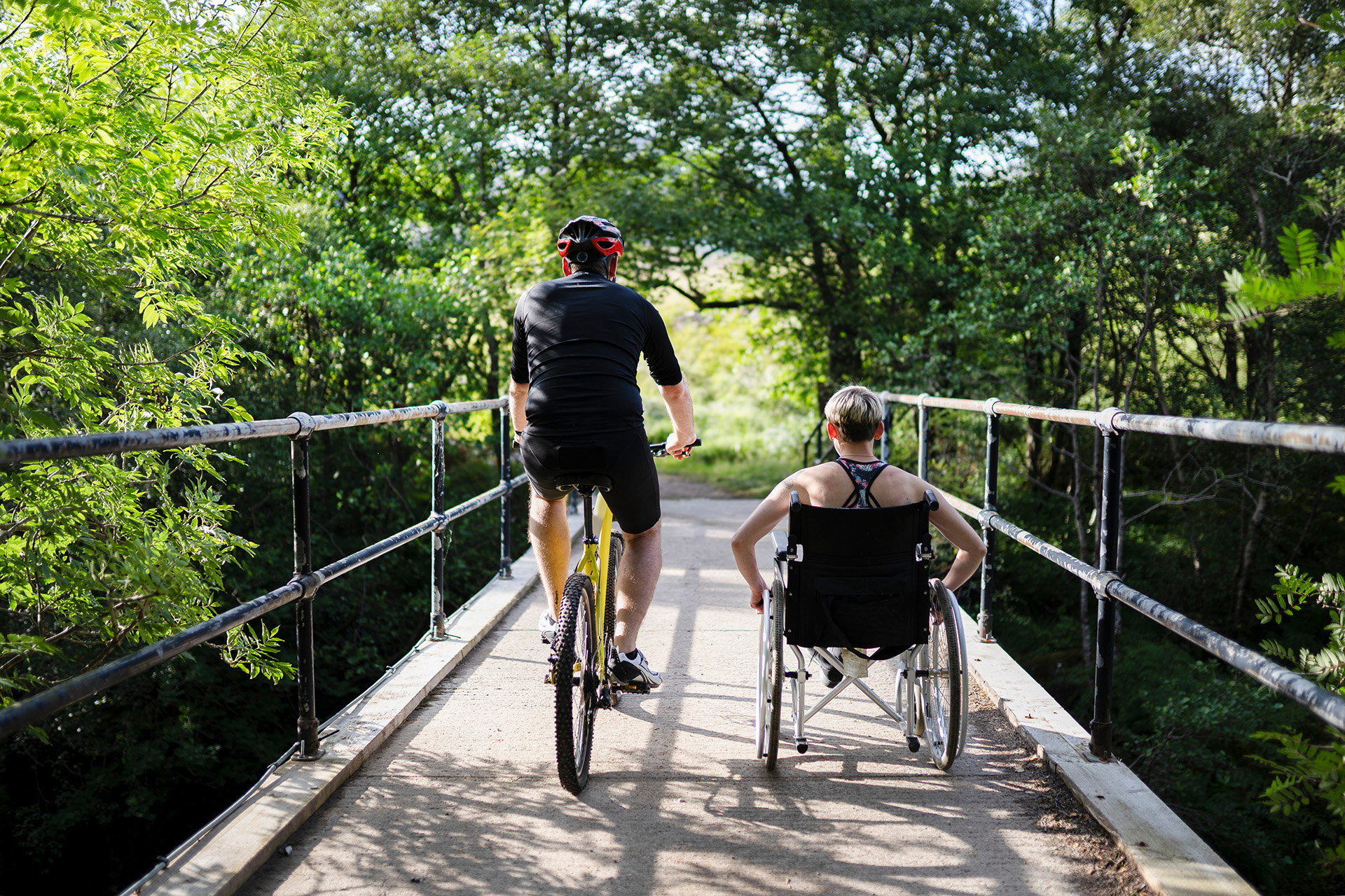 A man riding a bicycle in a park alongside a woman in a wheelchair.